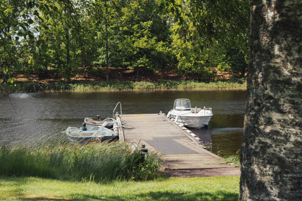 Wooden dock on a calm river with two small boats moored, surrounded by green trees and grass near Toftaholm Manor, with a tree trunk in the foreground.