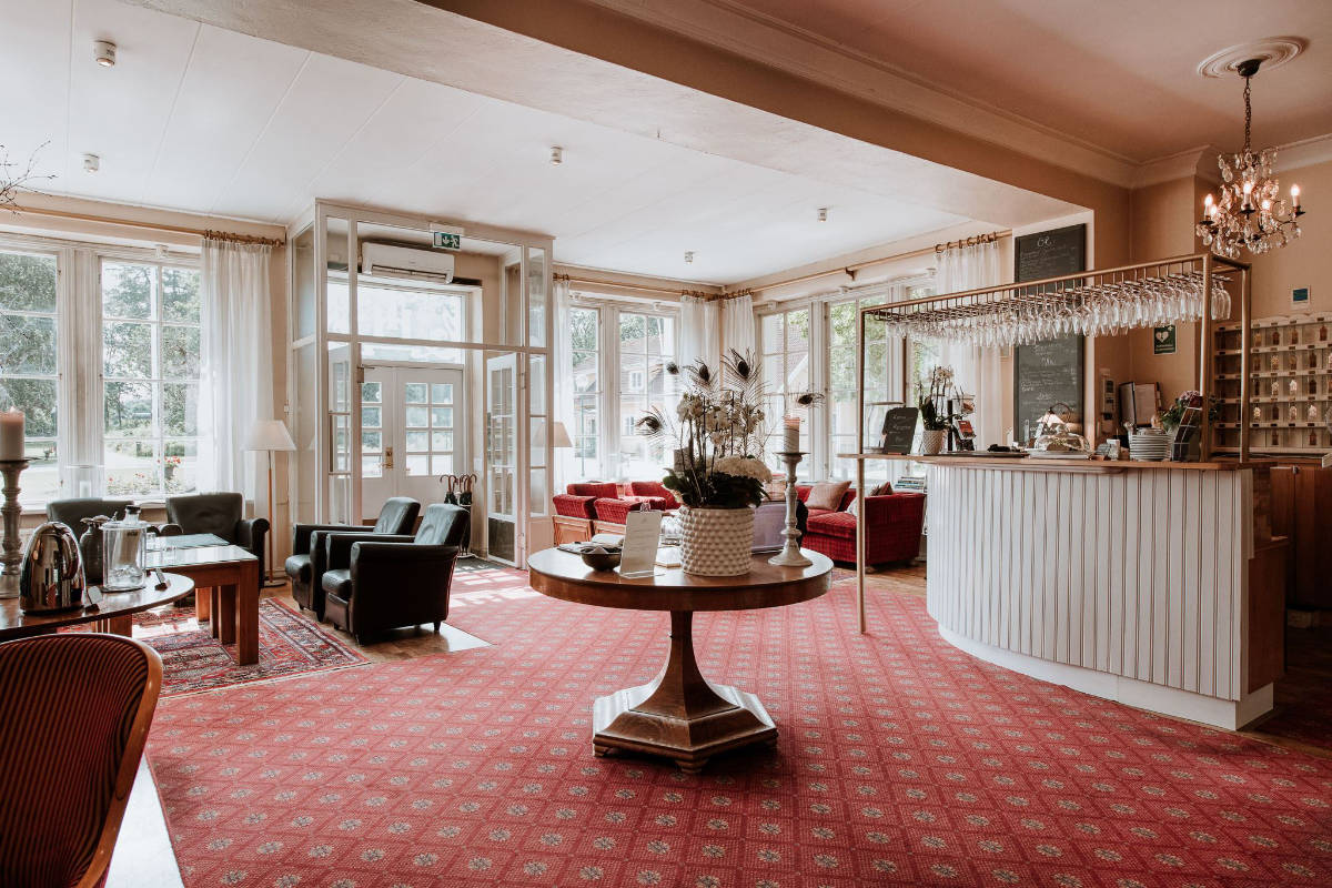 Spacious hotel lobby at Toftaholm Manor with red carpet, cozy seating areas, a round wooden table, and a white reception desk; large windows fill the space with natural light.