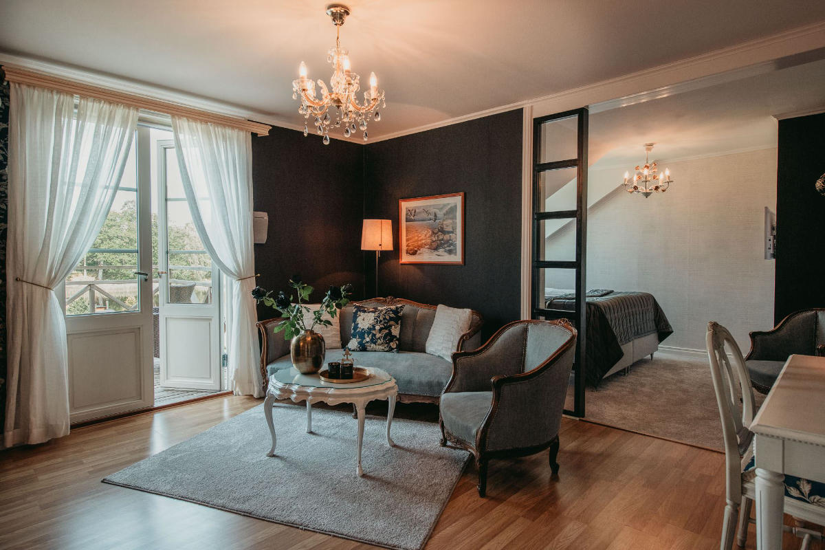 A stylish living area at Toftaholm Manor features a sofa, armchairs, coffee table, and chandelier, opening to a balcony; a bed is visible in a separate nook divided by a partial wall.