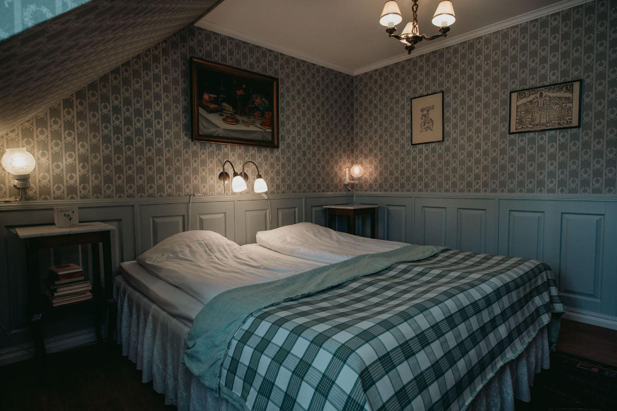 A cozy bedroom at Toftaholm Manor with two single beds pushed together, plaid bedding, patterned wallpaper, framed art, bedside tables, and soft ambient lighting.