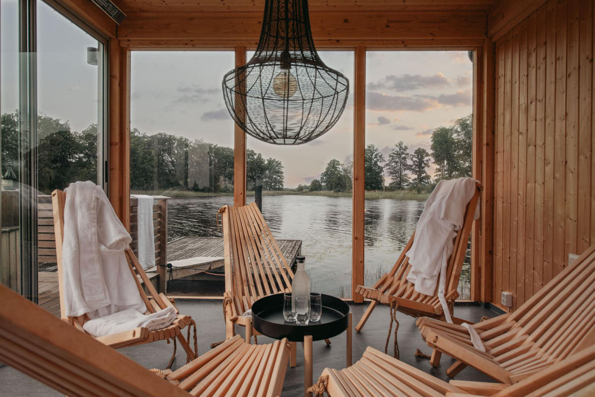 A wooden room at Toftaholm Manor features four lounge chairs, each draped with a white towel, surrounding a table with water bottles. Large windows offer a stunning view of the river and trees at sunset.