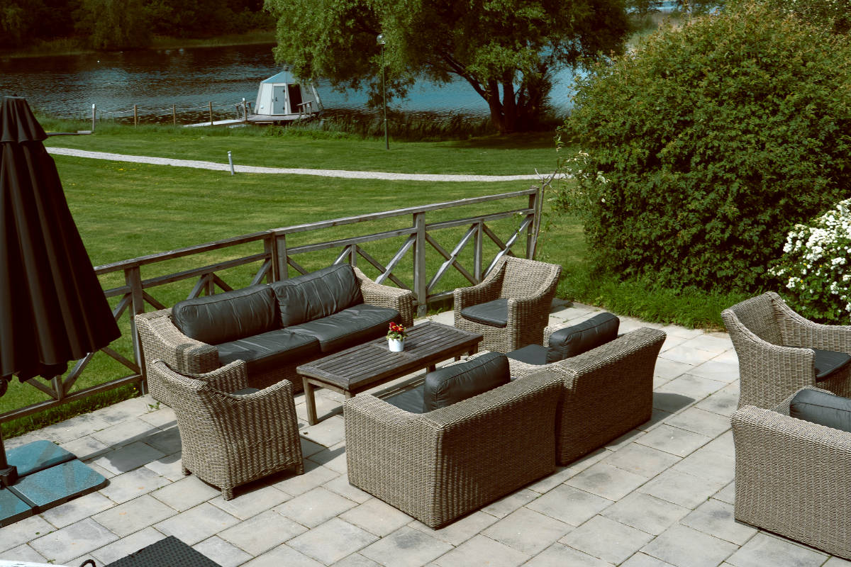 Outdoor patio with wicker furniture set, including sofas and armchairs around a table, overlooking a grassy area and a river with trees in the background at Toftaholm Manor.