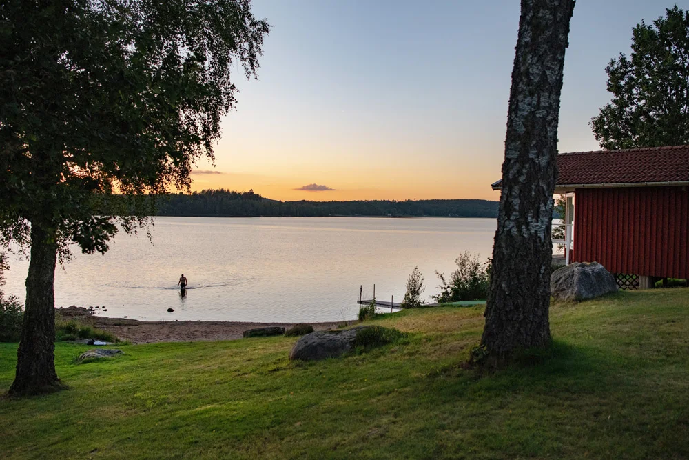 Une personne se tient dans une eau peu profonde près de la rive d'un lac au coucher du soleil, avec au premier plan des arbres, des rochers et l'emblématique bâtiment en bois rouge de Två Skyttlar.