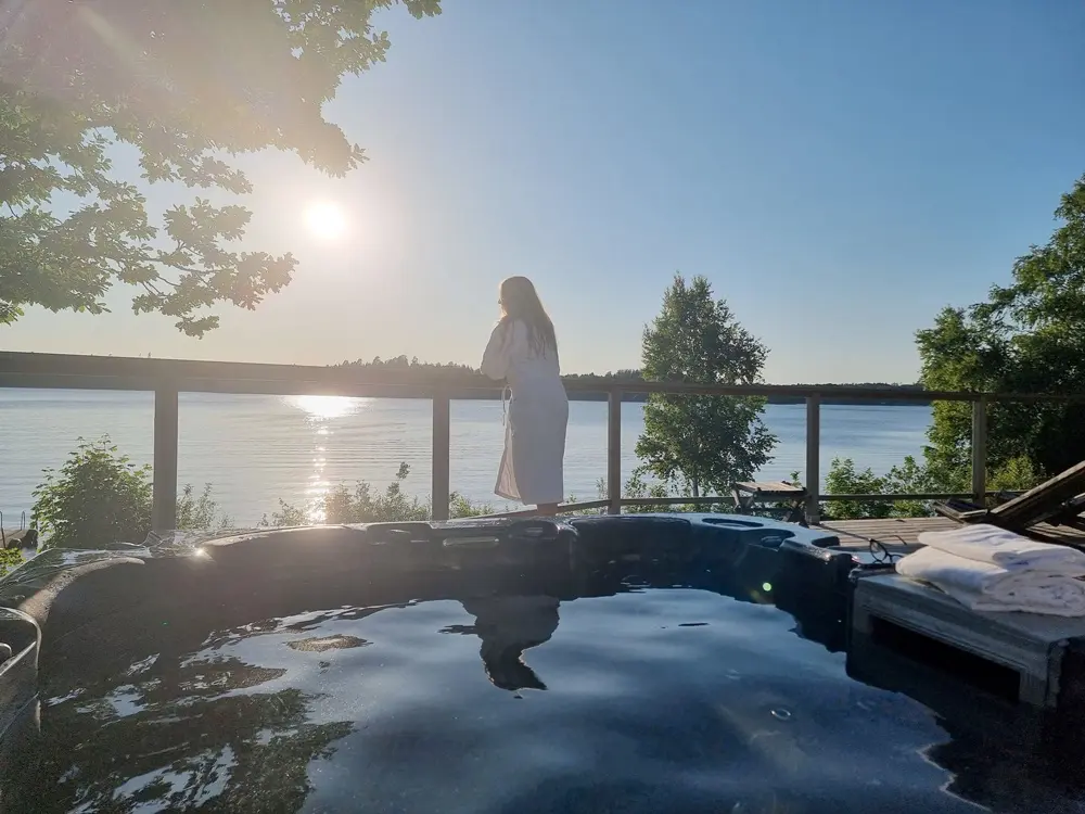 Une personne vêtue d'une robe blanche se tient sur la terrasse de Två Skyttlar, surplombant un lac au coucher du soleil, avec au premier plan un jacuzzi et des serviettes pliées.