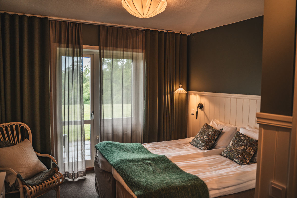 Une chambre d'hôtel Två Skyttlar confortable avec un lit double, des couvertures vertes, des oreillers décoratifs, une chaise en osier, des rideaux et une porte vitrée qui s'ouvre sur une vue de la verdure à l'extérieur.