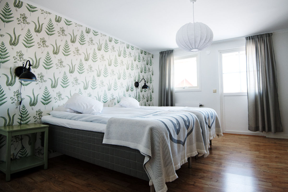 Une chambre lumineuse à Två Skyttlar avec deux lits simples poussés l'un contre l'autre, du papier peint à feuilles, deux lampes murales, une table de nuit verte, un sol en bois et une fenêtre avec des rideaux gris.