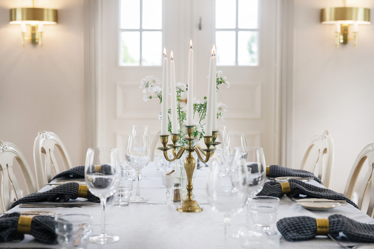 La table du Två Skyttlar est dressée avec des verres à vin, des serviettes noires et un chandelier doré avec des bougies et des fleurs blanches, dans une pièce lumineuse aux murs et fenêtres blancs.