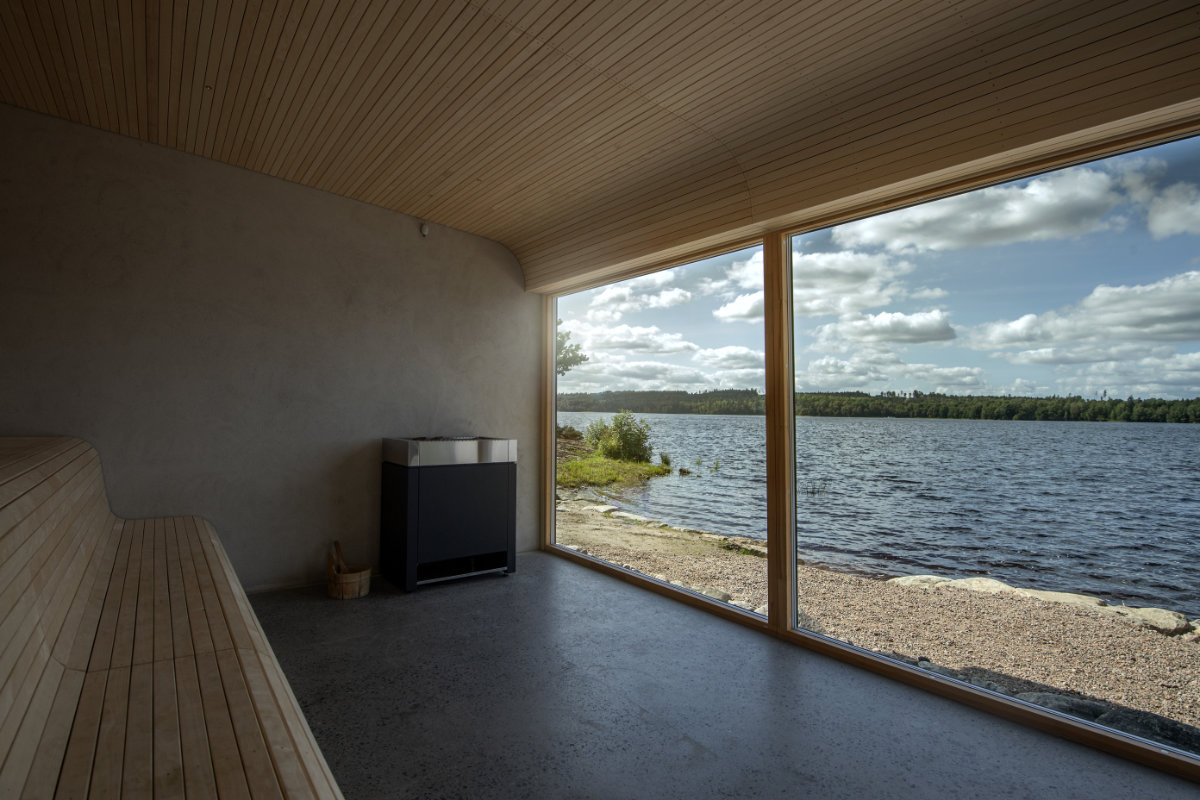 Le sauna moderne en bois du Två Skyttlar est doté d'une grande fenêtre allant du sol au plafond et donnant sur un lac serein et une forêt luxuriante sous un ciel partiellement nuageux.