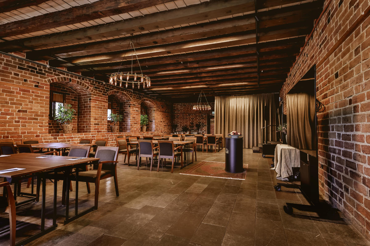 Une salle de réunion rustique au Vadstena Klosterhotel avec des murs en briques, des poutres en bois au plafond, des tables et des chaises en bois, des lustres et des plantes en pot sur les rebords des fenêtres.