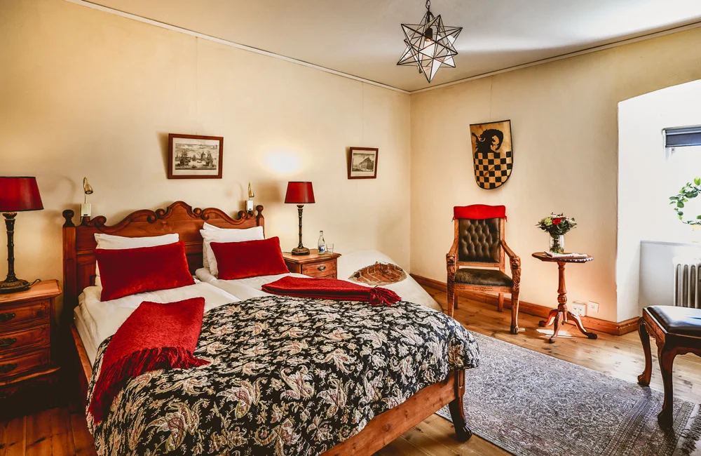 La chambre confortable du Vadstena Klosterhotel comprend un lit double avec des oreillers et une couverture rouges, deux tables de chevet avec des lampes, un fauteuil, une petite table ronde avec des fleurs et d'élégantes œuvres d'art murales.