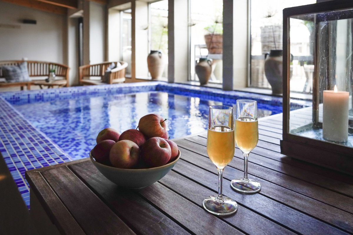 Un bol de pommes et deux coupes de champagne sont posés sur une table en bois à côté de la piscine intérieure au carrelage bleu du Vadstena Klosterhotel.