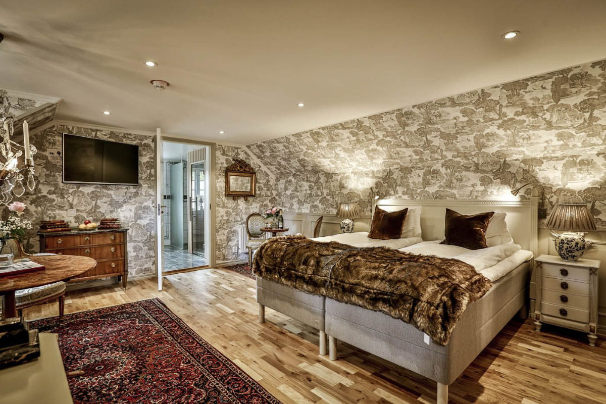 Une chambre confortable au Vadstena Klosterhotel avec du papier peint à motifs, des lits jumeaux avec des jetés de fourrure, un tapis rouge, des meubles anciens et une télévision murale.