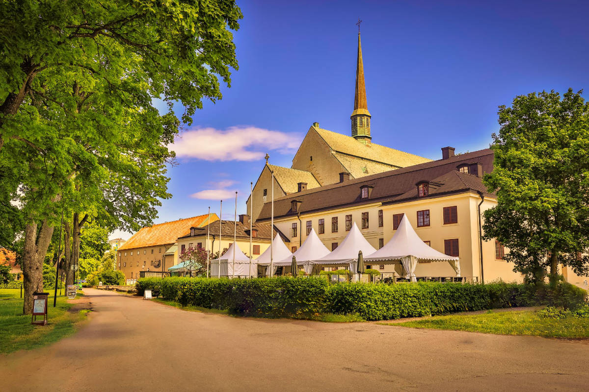 Un grand bâtiment historique avec un haut clocher, des tentes d'événement blanches devant, et un chemin bordé d'arbres sous un ciel bleu clair, rappelant l'élégant Vadstena Klosterhotel.