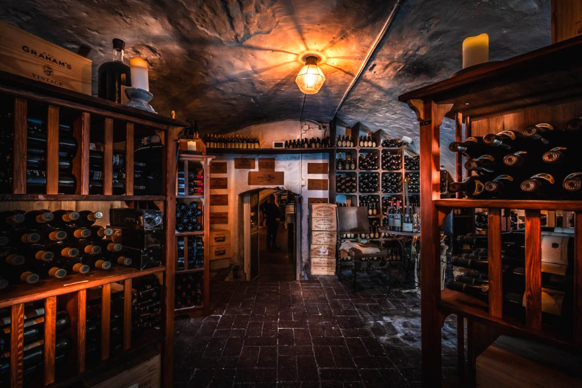 Une cave à vin faiblement éclairée au Vadstena Klosterhotel, avec des étagères remplies de bouteilles de vin, des caisses en bois et une porte centrale en pierre menant à une autre pièce.