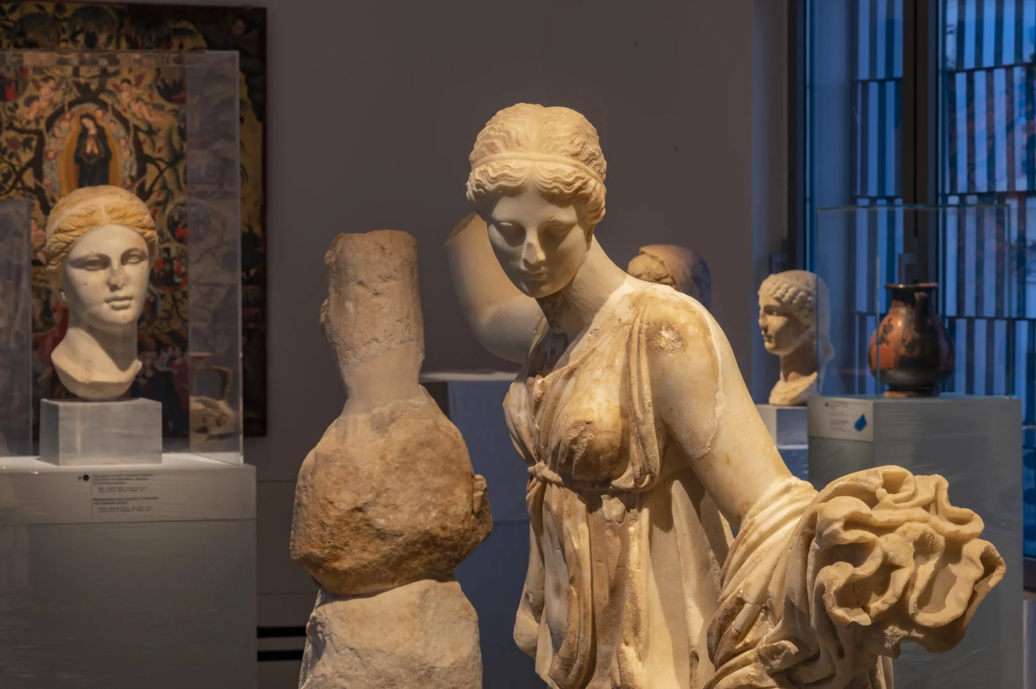 La magie de la fin de l'été à Santorin 2 Une statue de marbre représentant une femme se dresse dans une galerie de musée, évoquant la beauté sereine de Santorin à la fin de l'été, entourée de bustes anciens et d'objets classiques exposés dans des vitrines.