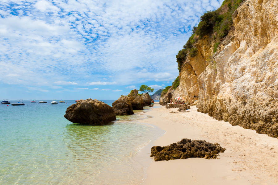 A sandy beach with clear turquoise water, rocky cliffs, and several boats anchored offshore under a partly cloudy sky.