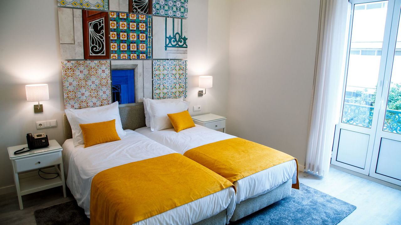 Habitación del Rio Art Hotel Setúbal con dos camas individuales, sábanas blancas y mantas amarillas, azulejos decorativos en la pared, mesillas de noche, lámparas y una ventana con cortinas transparentes.