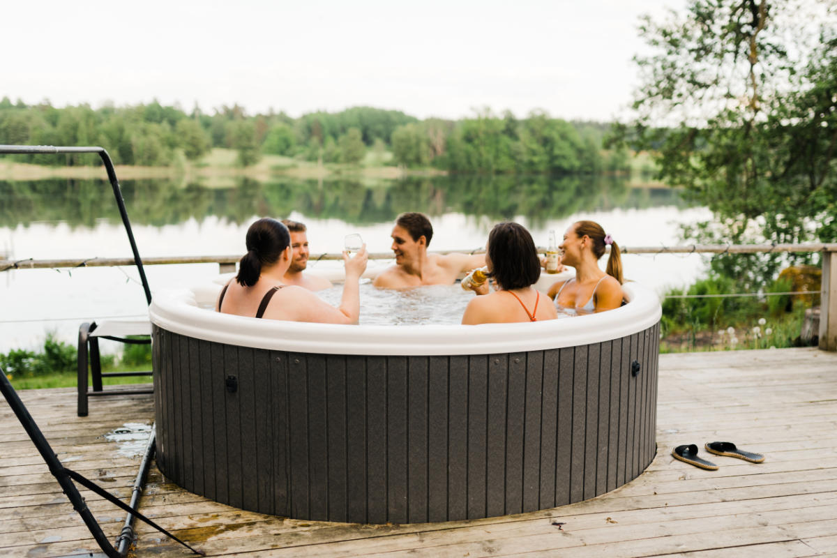 Quatre personnes se détendent dans un jacuzzi extérieur rond sur la terrasse en bois d'Asa Manor, en prenant un verre et en discutant, avec un beau lac et des arbres visibles en arrière-plan.