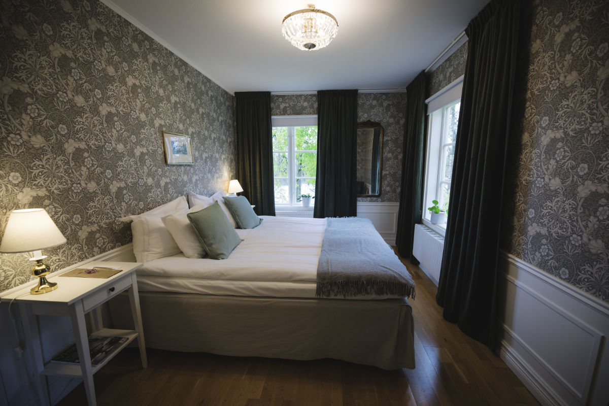 Un lit double bien fait avec des coussins verts trône dans une chambre à coucher du manoir d'Asa, avec du papier peint à fleurs, du parquet, un lustre, deux fenêtres et des tables de chevet avec des lampes.
