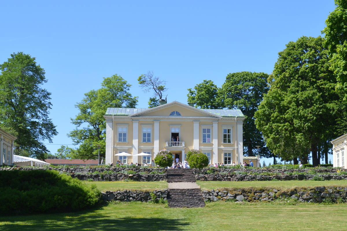Un manoir jaune de deux étages avec des colonnes blanches se dresse au milieu d'arbres verts, des marches en pierre y mènent et des personnes sont assises près de l'entrée sous un ciel bleu clair : c'est le charmant manoir d'Asa en Suède.