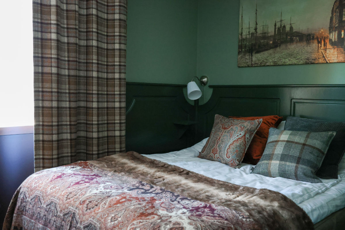 Un letto ben sistemato con cuscini a fantasia e una coperta paisley in una stanza dalle pareti verdi, con tende a quadri e un dipinto nautico sopra la testata.