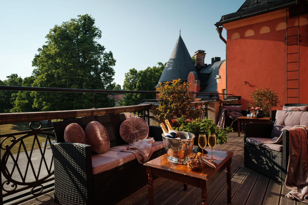 Terrazza all'aperto con cuscini, tavolo con champagne, bicchieri e stuzzichini, con vista sugli alberi e su un edificio storico con una torre a punta.