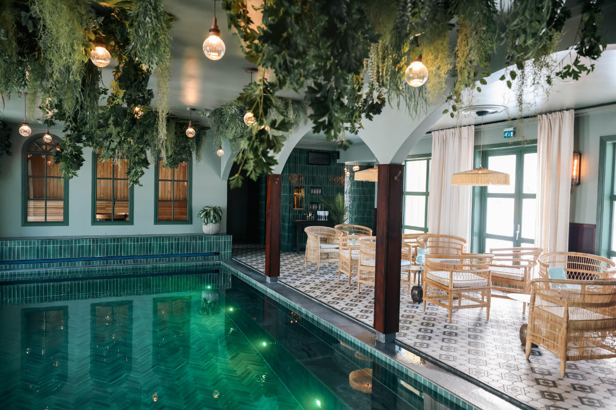 Area piscina interna con piastrelle verdi, piante pendenti, soffitti ad arco, sedie in vimini, pavimento piastrellato e luce naturale proveniente da grandi finestre con tende bianche.