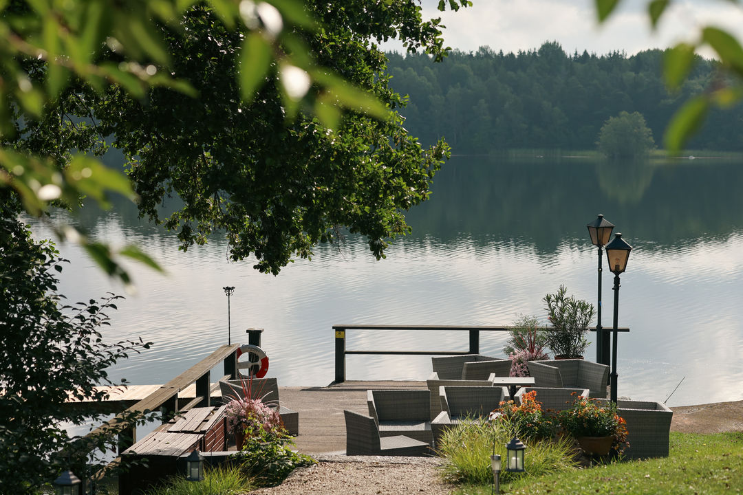 Area salotto all'aperto con sedie e tavoli in vimini su una terrazza in riva al lago, circondata dal verde e affacciata su acque tranquille con alberi sullo sfondo.