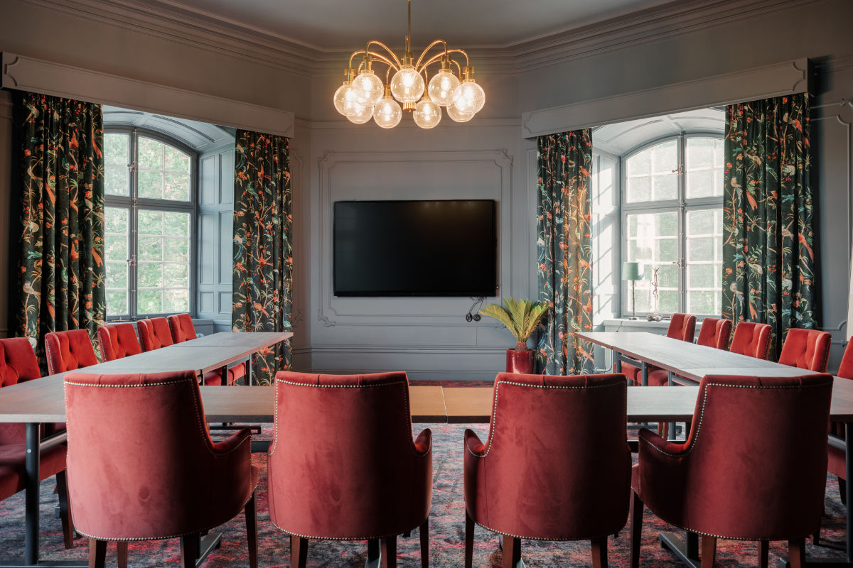 Una sala conferenze con sedie di velluto rosso, tavoli a U, un grande schermo a parete, lampadari e tende a fantasia sulle alte finestre.