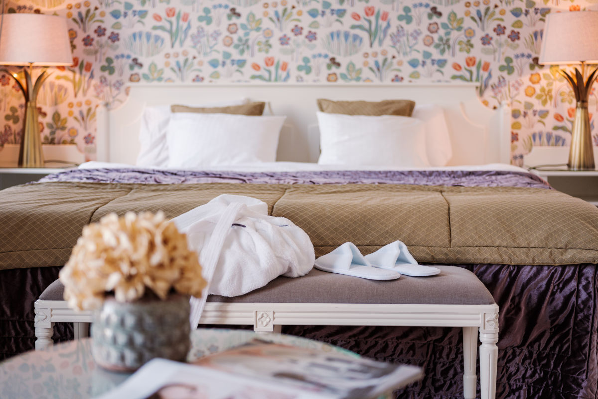 Camera d'albergo con letto ben fatto, carta da parati a fiori, due lampade, panca con accappatoio e ciabatte piegate, tavolo con composizione di fiori secchi e riviste.