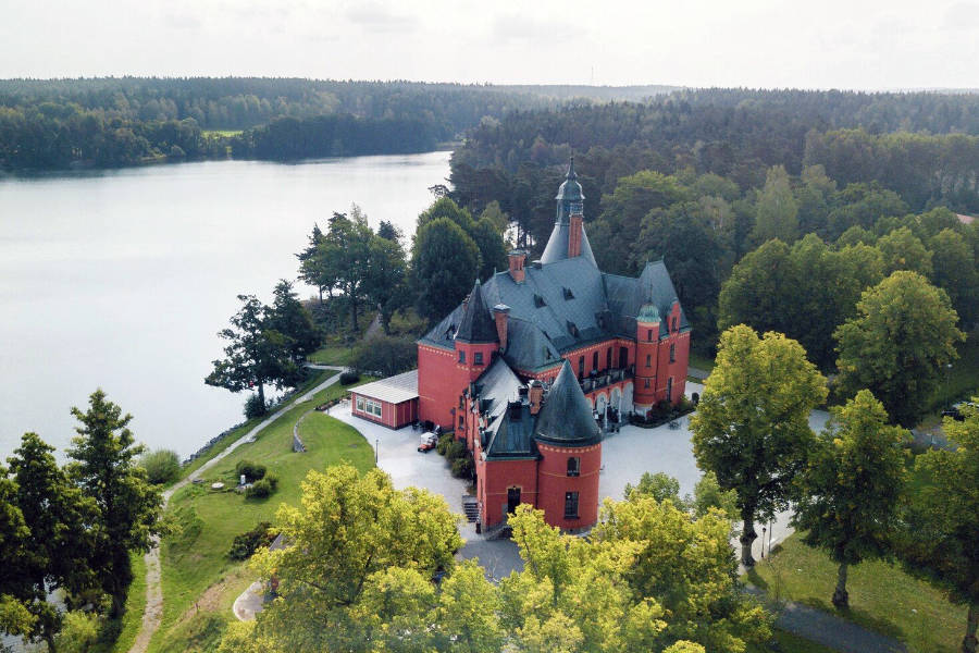 Aerial view of a large, red-brick castle with multiple turrets, surrounded by trees and located beside a lake.