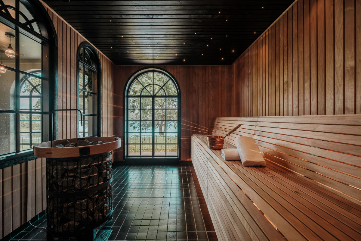 Una moderna sala sauna in legno con panche a più livelli, un secchio e un mestolo, grandi finestre ad arco e una stufa in pietra su un pavimento piastrellato.