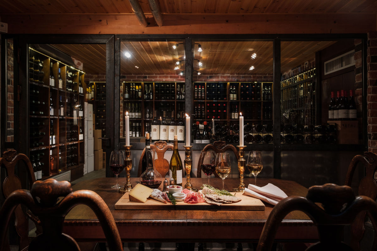 Una cantina poco illuminata con scaffali di bottiglie di vino, un tavolo di legno con bicchieri, candele, formaggio, pane e salumi.