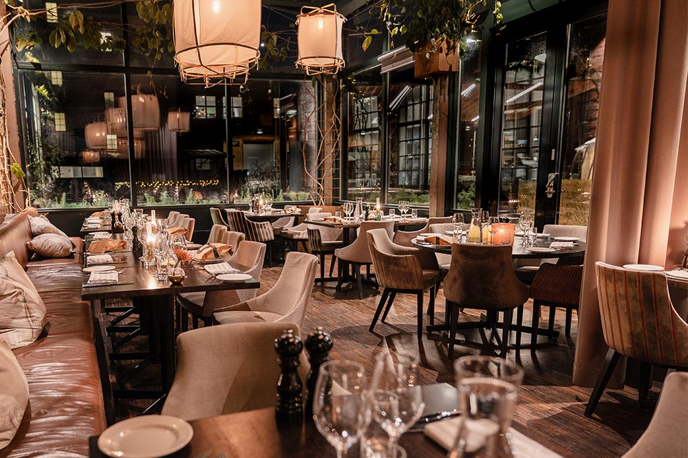 Ein warm beleuchteter, moderner Restaurantbereich mit gedeckten Tischen, gepolsterten Stühlen, Hängelampen und großen Fenstern mit Blick auf einen Garten.