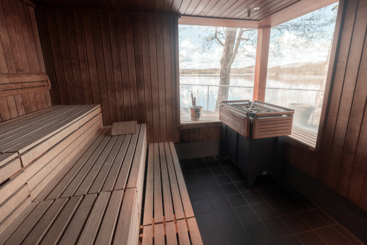 Eine hölzerne Sauna mit Bänken, einem Ofen, einem Wassereimer und großen Fenstern mit Blick auf einen See und Bäume draußen.