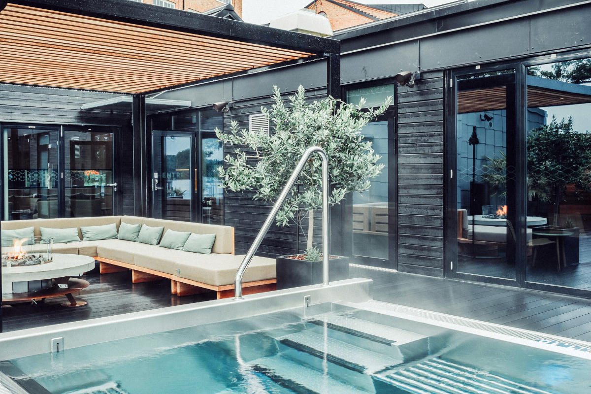 Moderne Außenterrasse mit einem dampfenden, rechteckigen Pool, einem hölzernen Sitzbereich mit Kissen, einem Topfbaum, einer Feuerstelle und Glastüren, die nach innen führen.