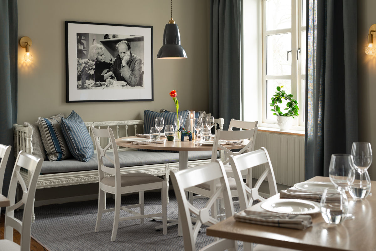L'accogliente sala da pranzo della Skytteholm Nordic Spa è caratterizzata da sedie bianche e tavoli rotondi apparecchiati per un pasto, una panca imbottita, una foto in bianco e nero incorniciata e una finestra che lascia entrare la luce naturale.