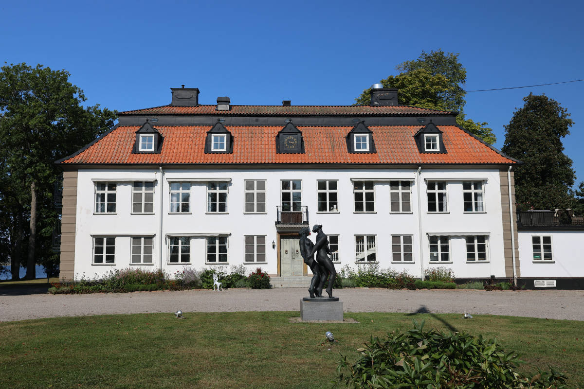 Vicino alla Skytteholm Nordic Spa si trova un edificio bianco a due piani con tetto a tegole rosse e finestre multiple, con una scultura nera di due figure posizionata sul prato di fronte.