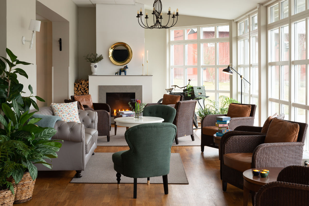Un accogliente soggiorno ispirato alla Skytteholm Nordic Spa, con poltrone e un divano attorno a un tavolino bianco, un camino, ampie finestre e varie piante e oggetti decorativi per un rifugio rilassante.
