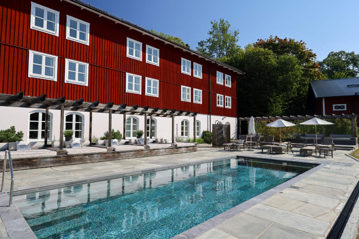 Piscina all'aperto con sedie a sdraio e ombrelloni accanto all'edificio rosso a più piani della Skytteholm Nordic Spa con finiture bianche in una giornata di sole.