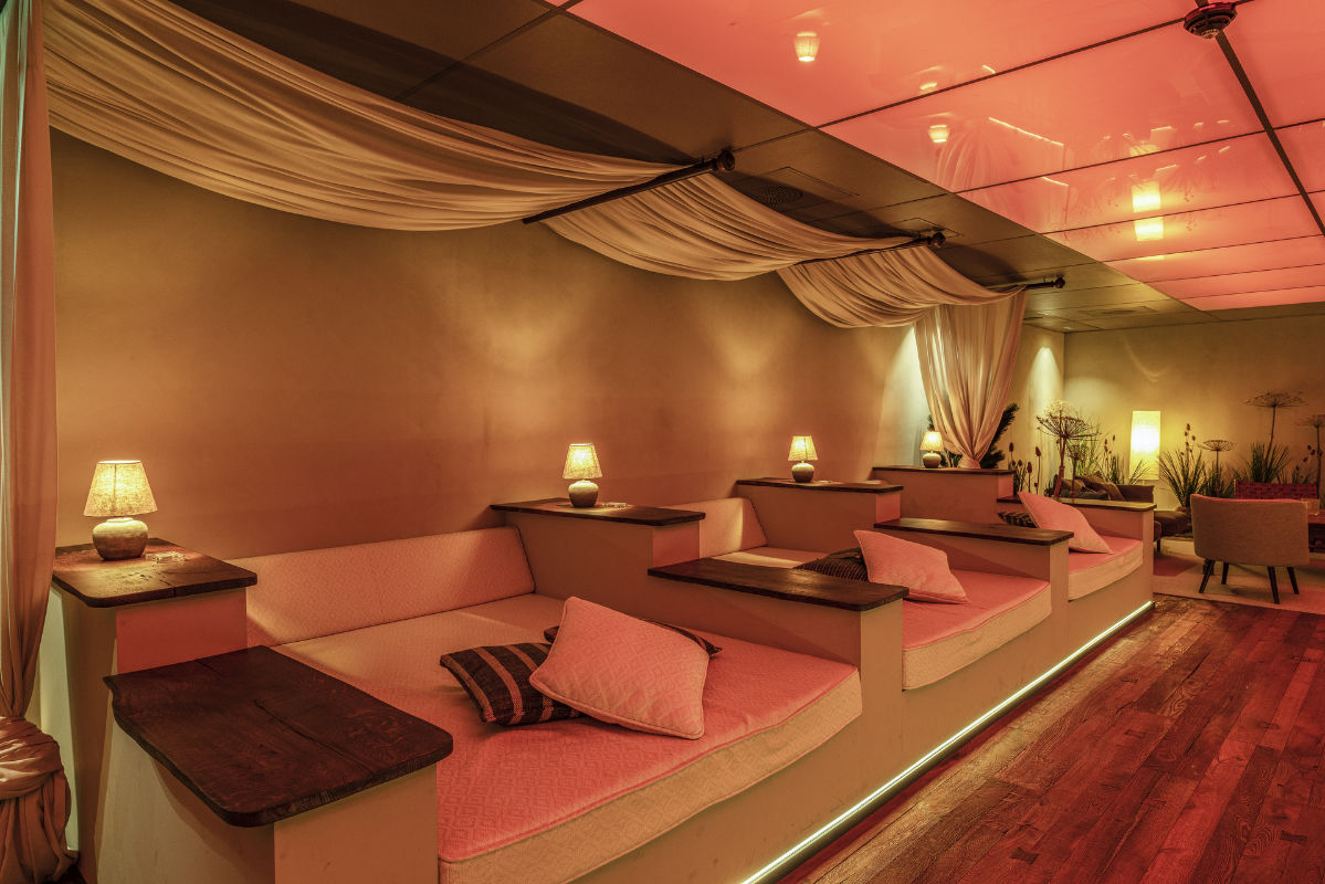 Una fila di salottini privati della Skytteholm Nordic Spa è caratterizzata da cuscini rosa, piccole lampade su tavolini di legno e tessuti drappeggiati in alto sotto una calda illuminazione rossa.