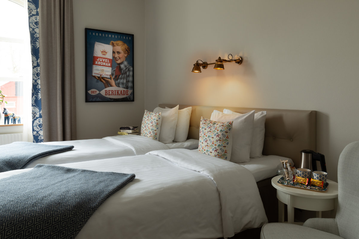 Una camera d'albergo della Skytteholm Nordic Spa con due letti singoli, lenzuola bianche, cuscini a fantasia, opere d'arte alle pareti, lampade da comodino e un vassoio con snack, bollitore e tazze su un tavolino.