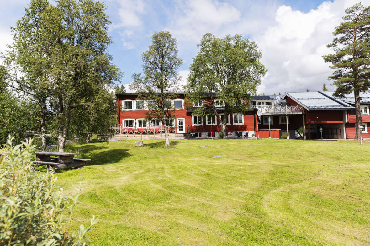 Le Storsätra Mountain Hotel est un grand bâtiment rouge avec de nombreuses fenêtres et balcons, situé sur une pelouse soigneusement tondue et entouré de grands arbres sous un ciel partiellement nuageux.