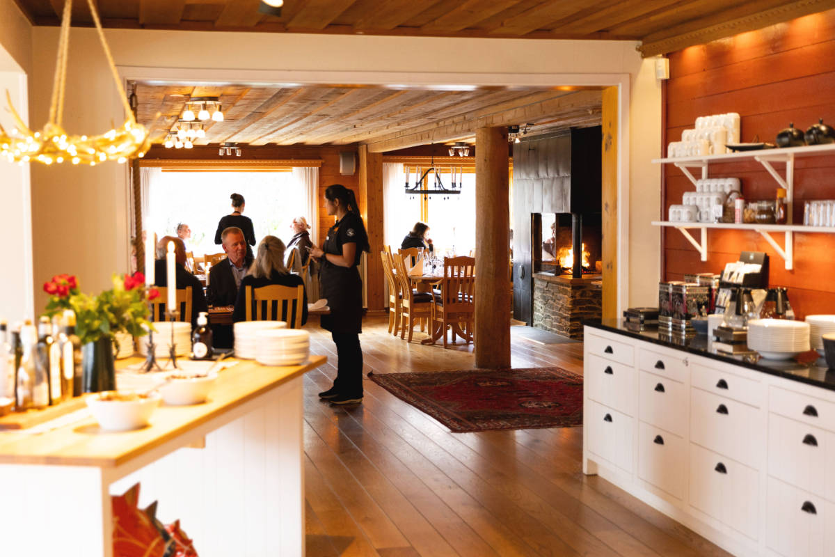 Une serveuse sert les clients aux tables d'un restaurant confortable et chaleureusement éclairé, doté de parquet et d'une cheminée, au Storsätra Mountain Hotel, où des étagères présentent des plats et des verres bien rangés.