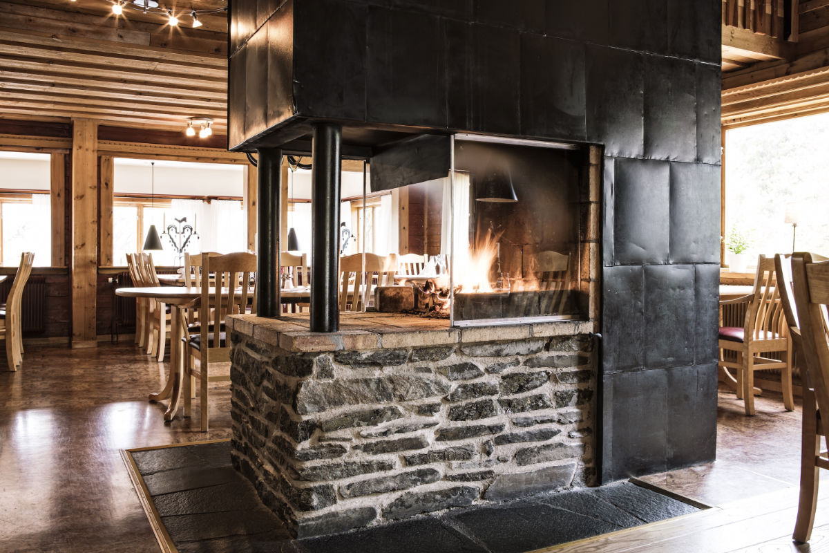 Une cheminée en pierre et en métal est au centre de la salle à manger rustique du Storsätra Mountain Hotel, avec des chaises et des tables en bois et des poutres apparentes.