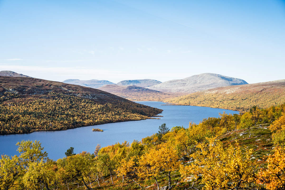 Un lac entouré de collines et d'arbres d'automne sous un ciel bleu clair. L'hôtel de montagne Storsätra, situé à proximité, offre une vue imprenable sur ce paysage serein.