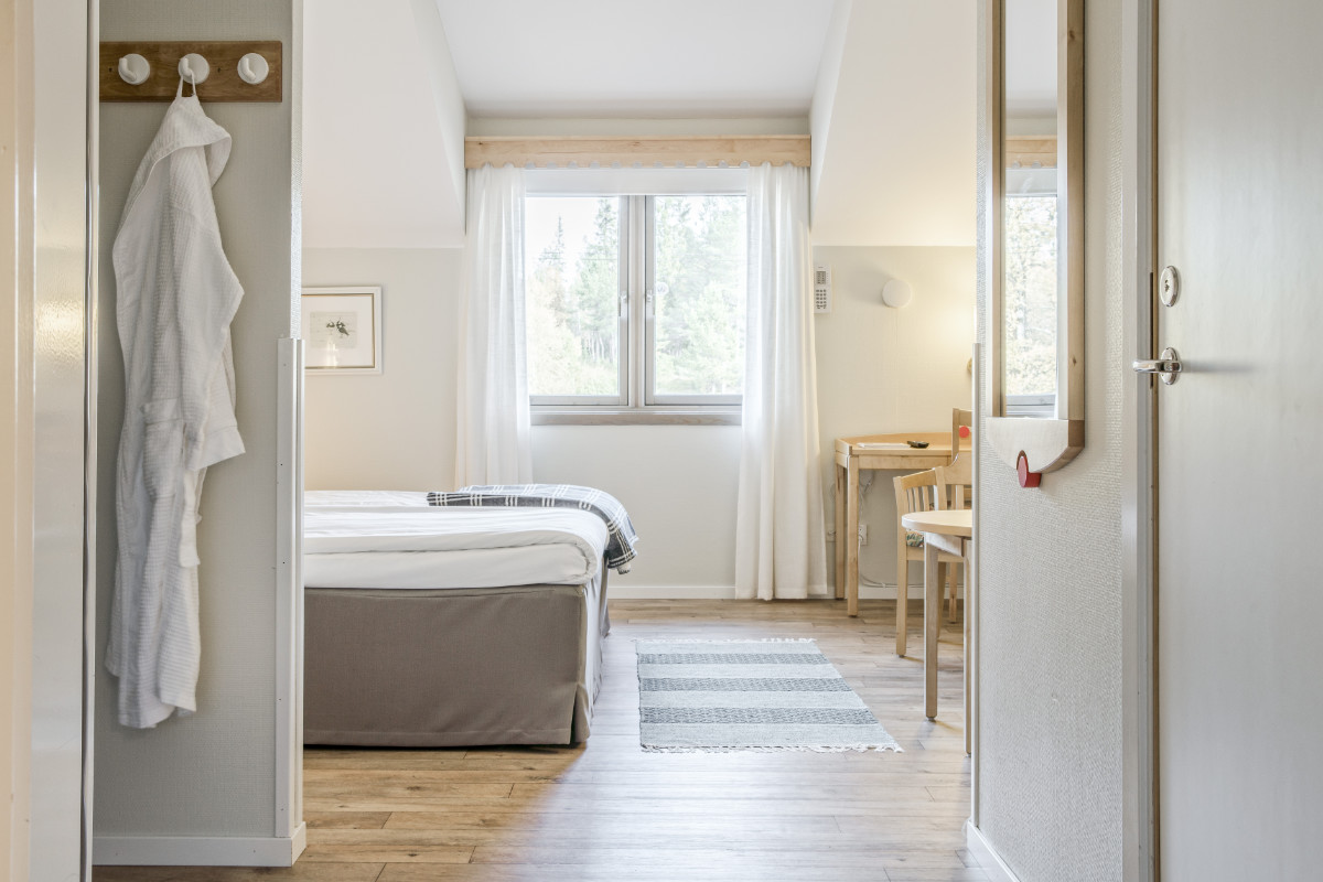 Les chambres claires et bien rangées du Storsätra Mountain Hotel disposent d'un lit double, de parquet, d'une table avec deux chaises, de rideaux blancs et d'un peignoir accroché à un crochet près de la porte.