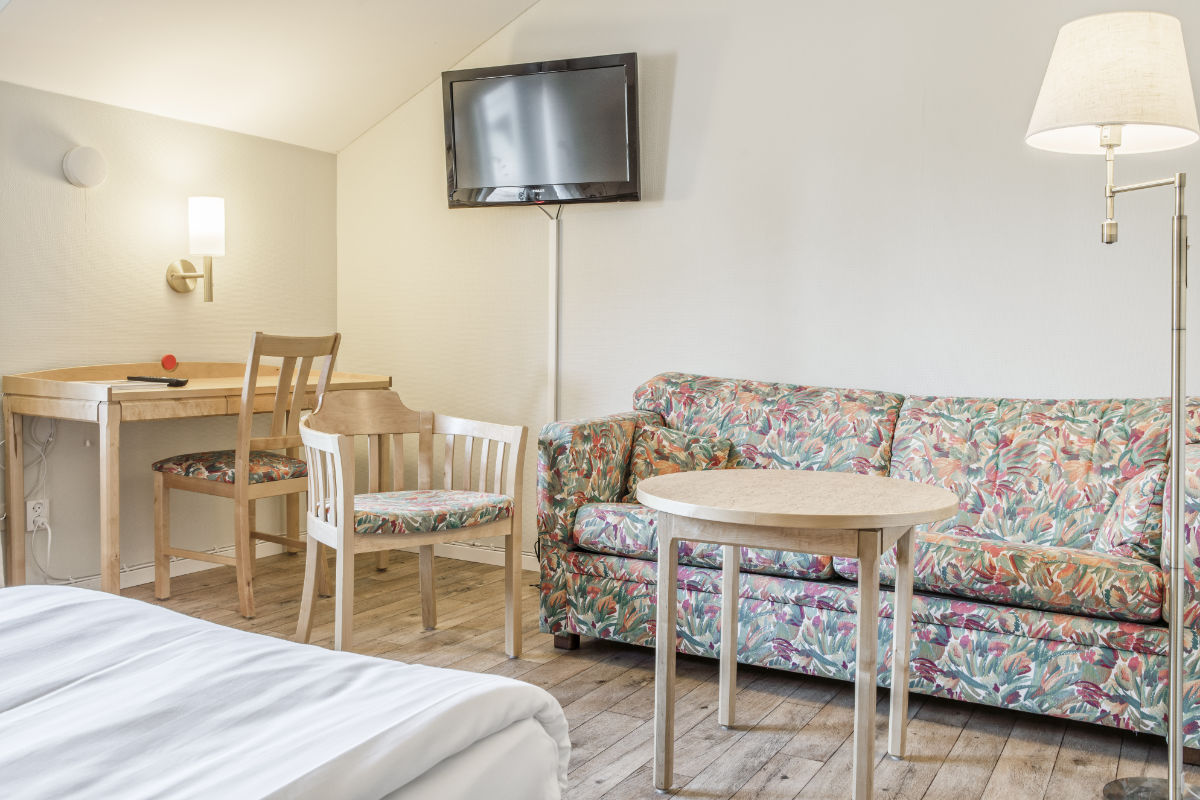 La chambre confortable du Storsätra Mountain Hotel dispose d'un canapé à motifs floraux, de chaises en bois, d'une table ronde, d'un bureau avec une lampe et d'une télévision murale sur un élégant sol en bois.