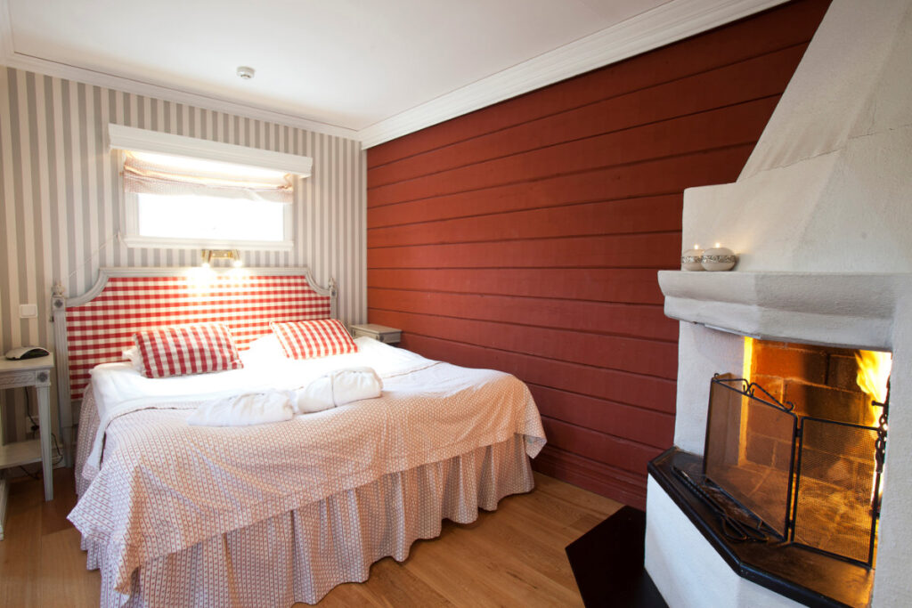 Belle come un quadro: cinque destinazioni europee che ispirano gli artisti 15 Un'accogliente camera da letto con letto matrimoniale, biancheria da letto a scacchi rossi e bianchi, carta da parati a righe, un caminetto acceso e una finestra che fa entrare la luce naturale.