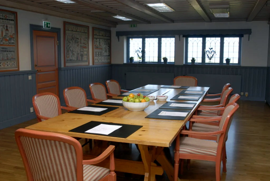 A long wooden conference table with striped chairs, placemats, documents, and a bowl of fruit in a room with decorative walls and windows.